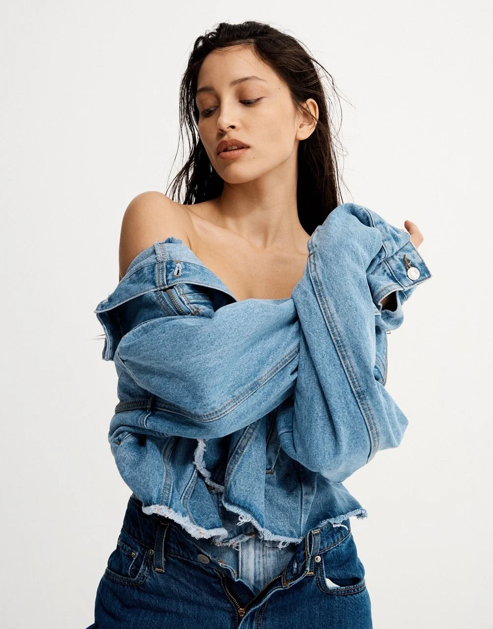 Young woman with long dark hair wearing a distressed denim jacket off her shoulders and jeans, posing against a plain white background.