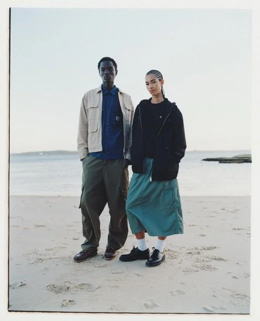 Two young adults standing on the beach with ocean and sky in the background.