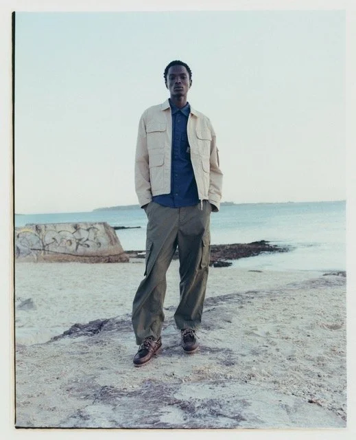A person standing on a beach with sand, rocks, and a body of water in the background.