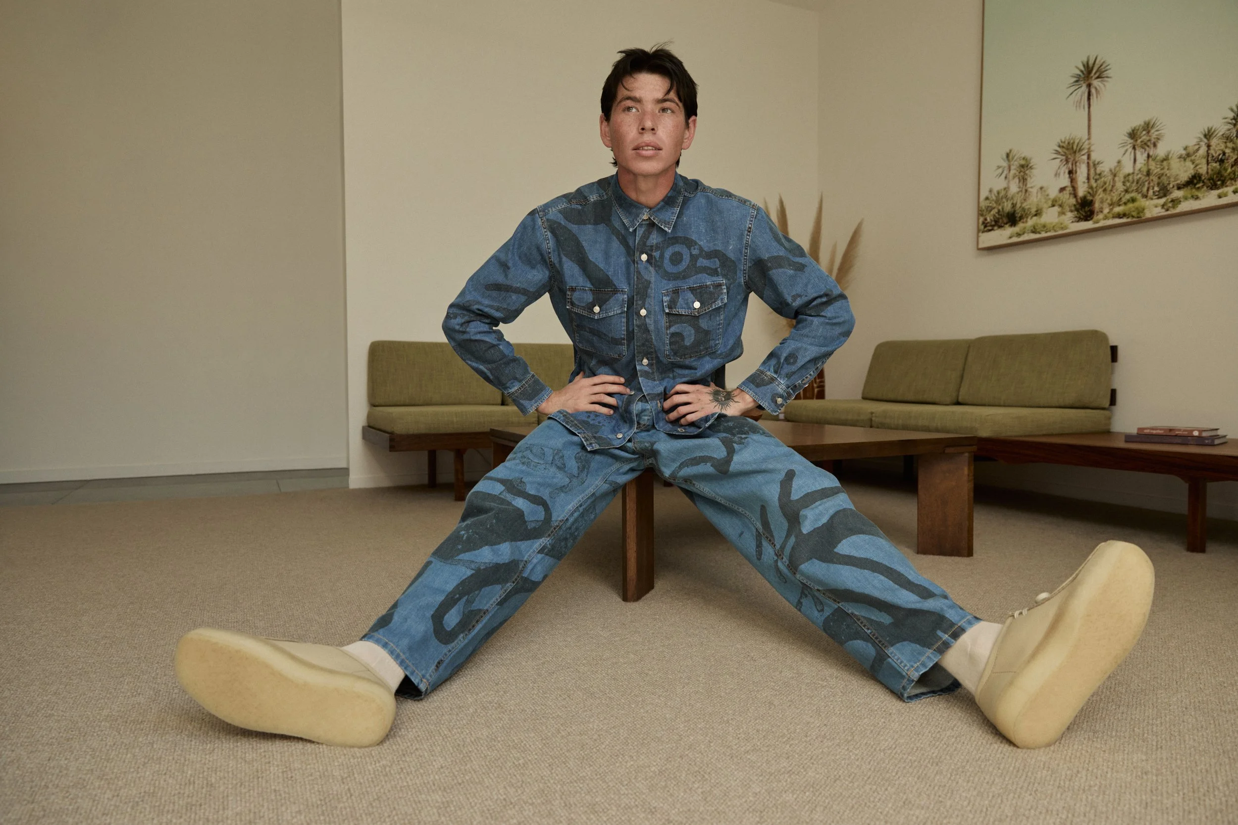 A person with short dark hair and tattoos on their hands and wrist, wearing a matching blue camouflage denim jacket and pants, sitting with legs wide apart on the floor in front of a bench inside a room with beige carpet and walls, a painting of dese