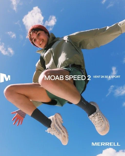 Person jumping in the air outdoors against blue sky, wearing outdoor gear with Merrell branding