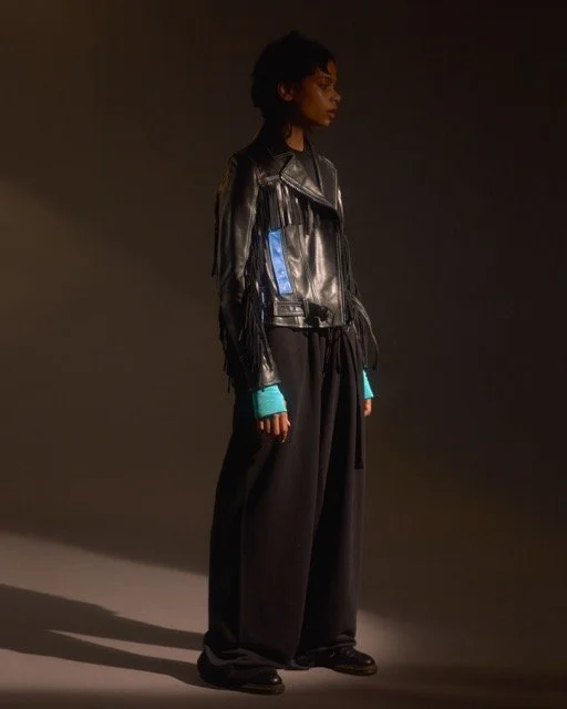 A woman with dark curly hair wearing a black leather jacket, teal fingerless gloves, and loose-fitting black pants against a dark background.