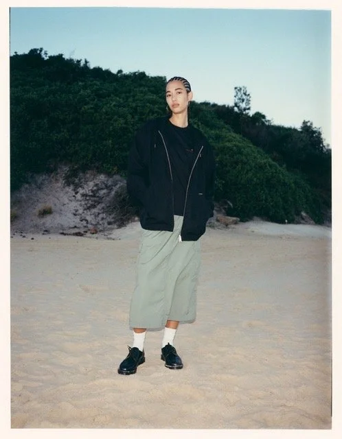 A young woman standing on a sandy beach with green bushes and a clear sky in the background. She is wearing a black jacket, beige wide-leg pants, white socks, and black shoes, with her hair styled in cornrows.