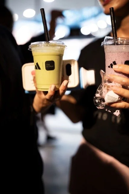 Two people holding colorful bubble tea drinks with black straws, one with a green drink and the other with a pink drink, in a busy indoor setting.