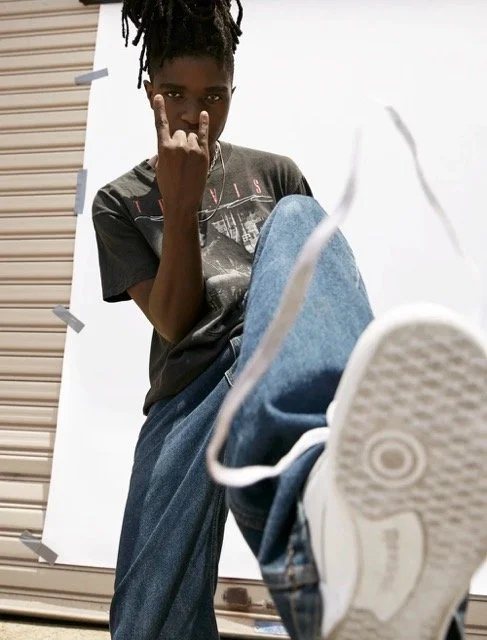 Person with dreadlocks wearing a dark T-shirt and jeans, making a rock gesture with their hand, and sitting with one leg extended toward the camera.