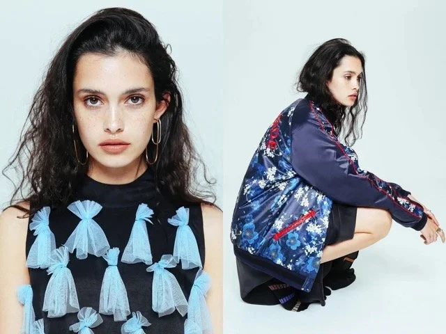 A young woman with dark, curly hair is looking at the camera, wearing a sleeveless black top with blue ribbon bows, and hoop earrings. Beside her, in a separate pose, she is squatting in profile, wearing a shiny blue floral jacket over black clothing