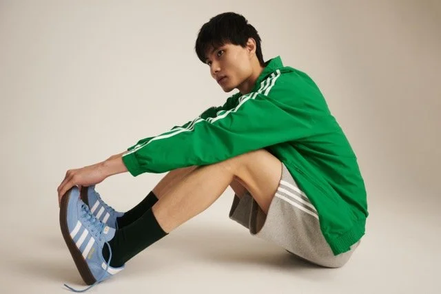 Young man sitting on the floor, wearing a green jacket, gray shorts, black socks, and blue sneakers, with a neutral background.