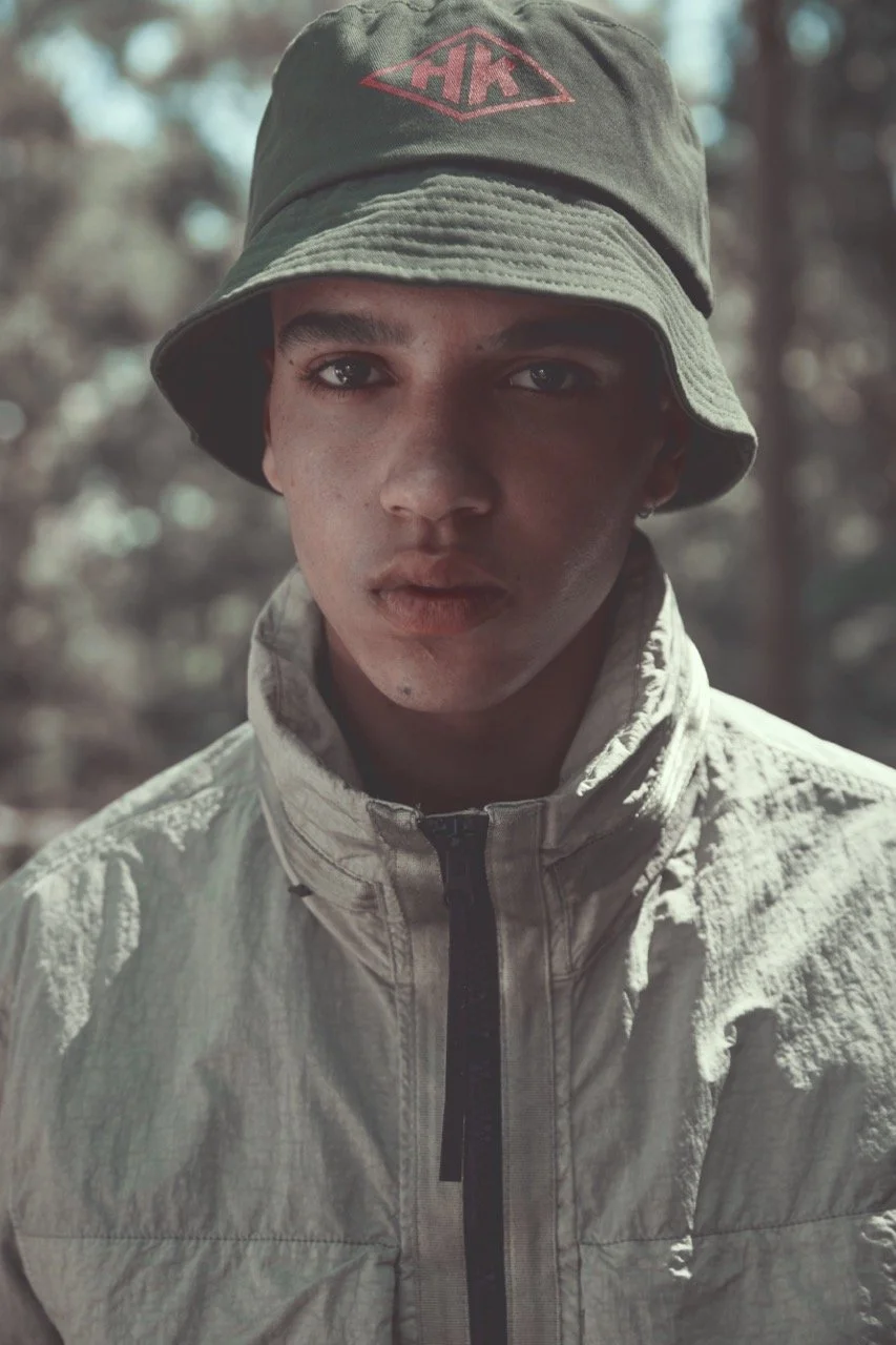 A young man wearing a bucket hat with a logo and a light-colored, quilted jacket standing outdoors in a wooded area.