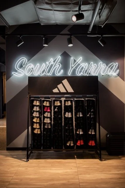 Neon sign reading "South Yanna" above a rack of folded athletic shoes, with a black Adidas logo on the wall behind.