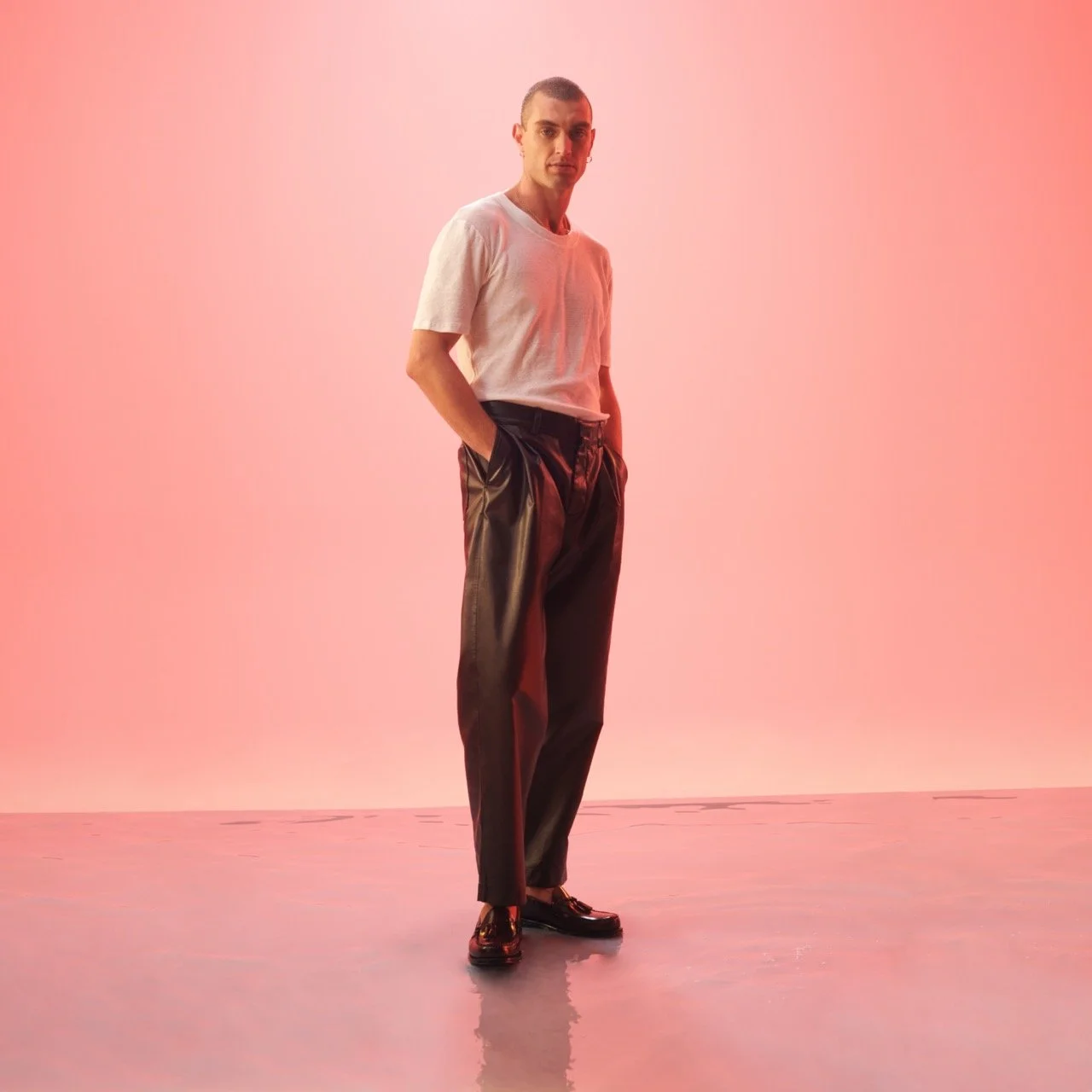 A young man standing against a pink gradient background, wearing a white t-shirt, black shiny pants, and black shoes, with hands in pockets and looking at the camera.