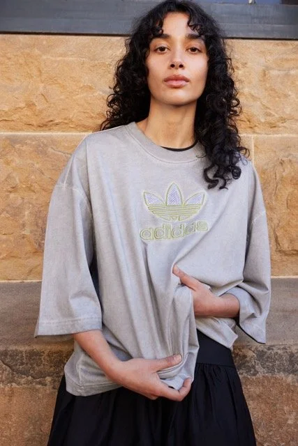 Young woman with curly dark hair wearing a loose gray Adidas t-shirt with the Adidas logo and black pants, standing outdoors against a brick wall.