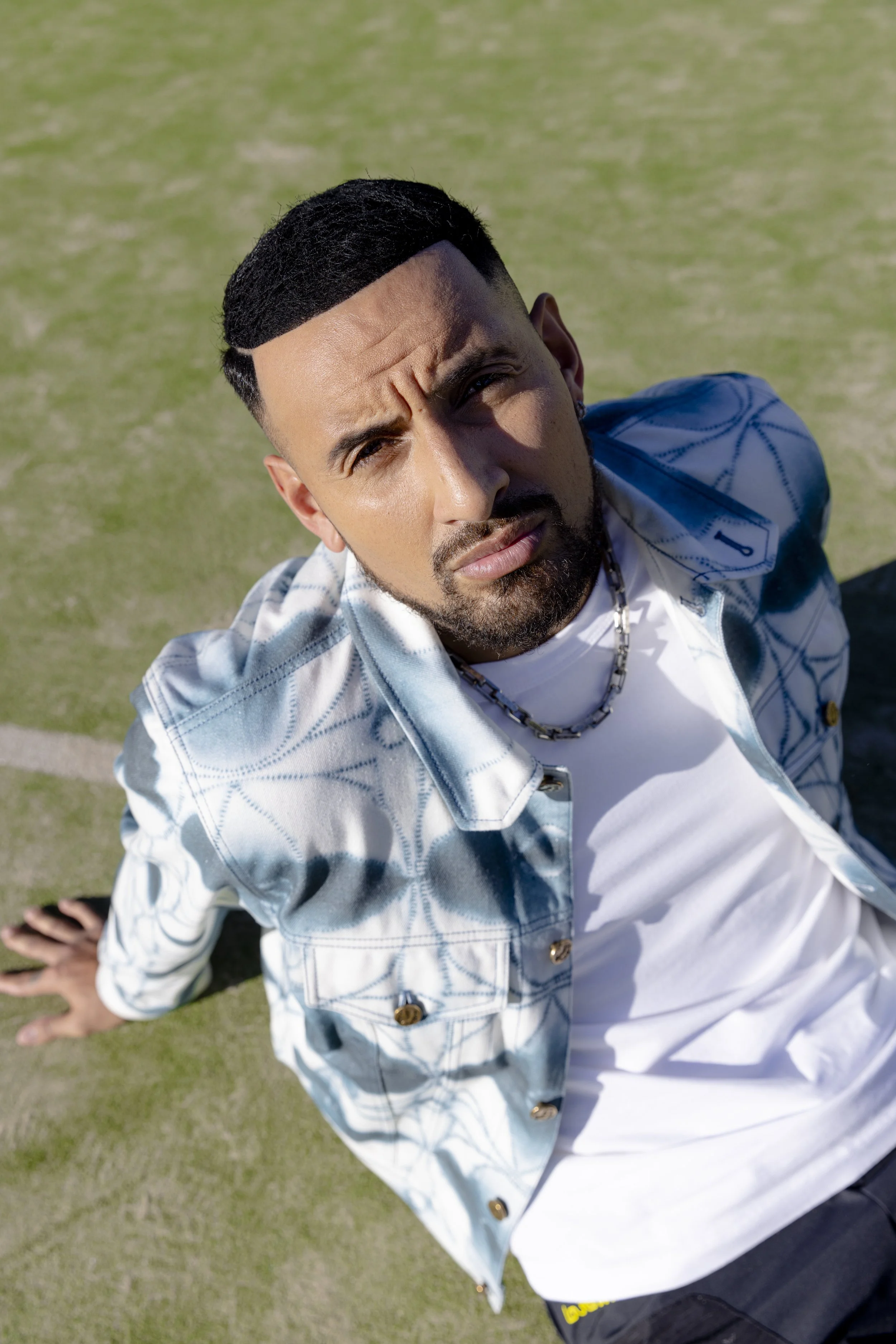 A man with dark hair and beard taking a selfie from a high angle outdoors on a grassy field. He is wearing a blue and white patterned denim jacket over a white shirt, with a silver chain necklace, leaning back on one hand.