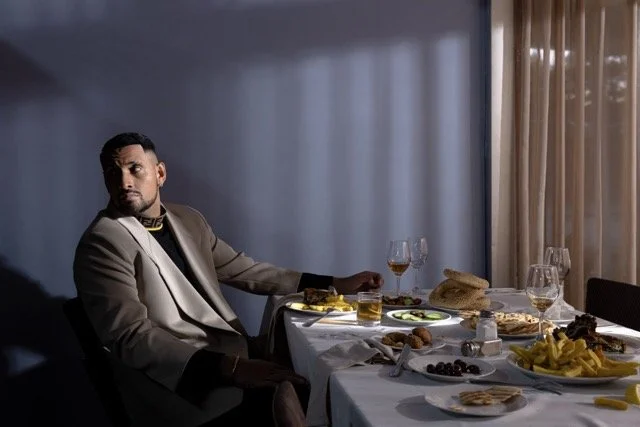 A man in a beige suit sitting at a dining table with food and drinks, in a dimly lit room with sheer curtains.
