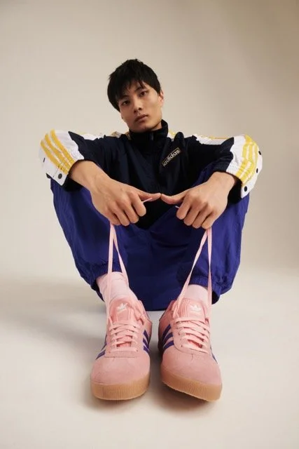 Young man sitting with legs crossed, holding pink sneakers in front of him, against a plain beige background.