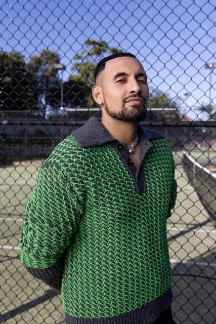 A man with dark hair and a beard standing in front of a chain-link fence at a tennis court outdoors, wearing a green patterned sweater and a gray collar.