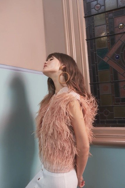 Young woman with brown hair and hoop earrings standing indoors near a stained glass window, wearing a pink feathered top and white pants.
