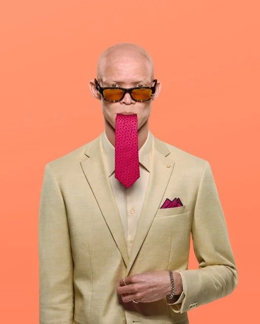 An elderly man with glasses and a pink tie hanging from his mouth against a peach background.