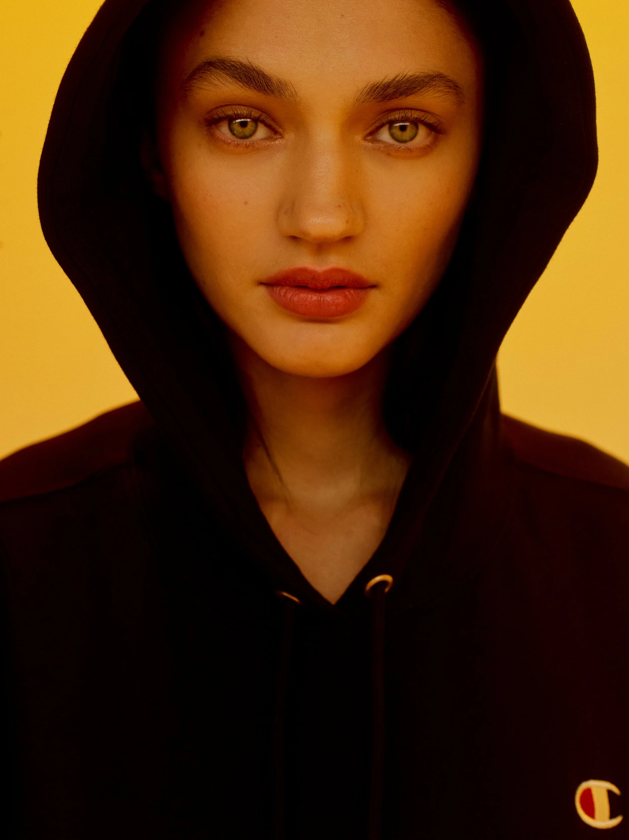Close-up image of a young woman with light complexion, hazel eyes, dark eyebrows, and dark hair, wearing a black hoodie with a yellow logo and hood up, against a yellow background.