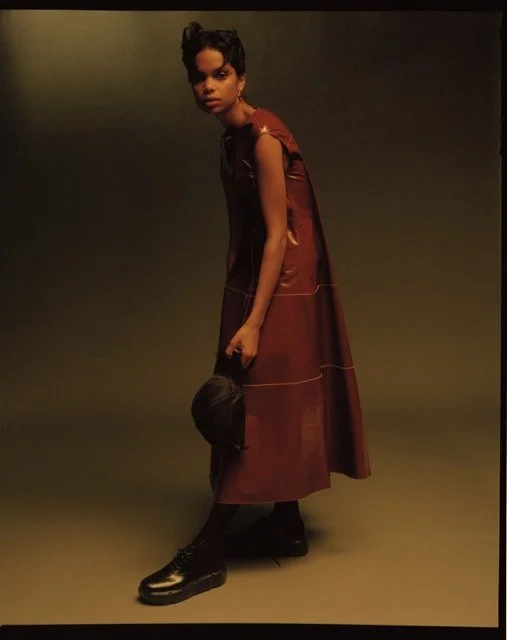 A woman with short hair wearing a long, sleeveless brown dress, black boots, and holding a black bag, standing against a dark background.