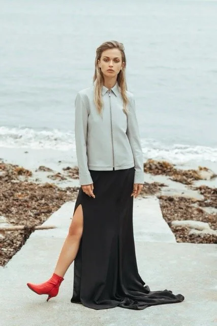 A woman standing on a concrete pathway near the ocean, wearing a light gray zip-up jacket, a long black skirt with a high slit, and red ankle boots, with a serious expression and wavy blonde hair.