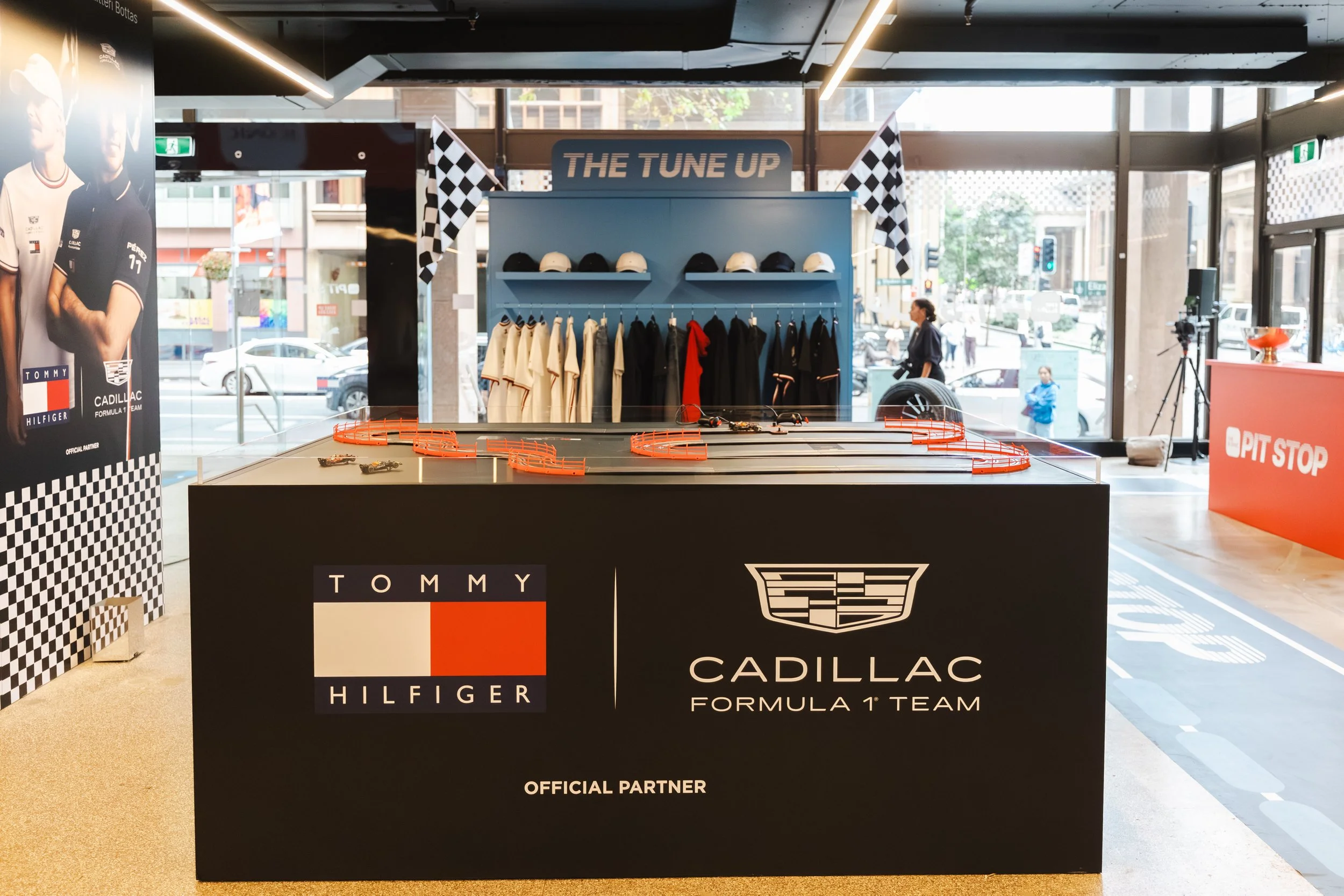 Display table with Tommy Hilfiger and Cadillac logos, featuring helmets, racing suits, and toy race cars, in a retail store area.