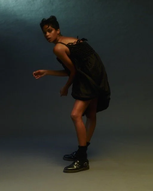 A woman with short dark hair wearing a black dress and black shoes standing in a dark studio.