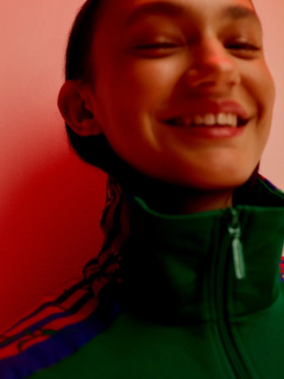 Smiling young boy wearing a green jacket with red and black stripes, standing against a pink wall.
