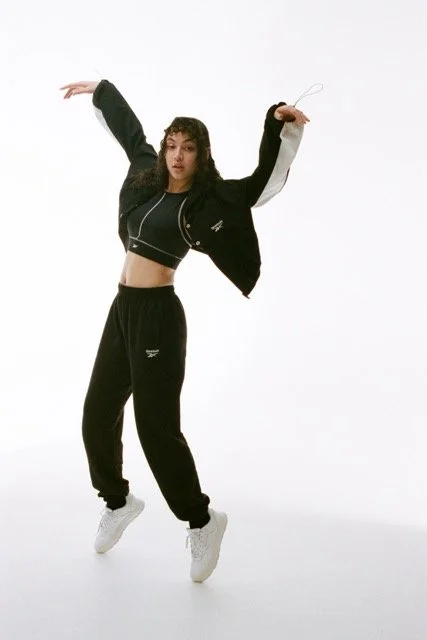 Young woman dancing in a black and white jacket, black sweatpants, and white sneakers against a white background.