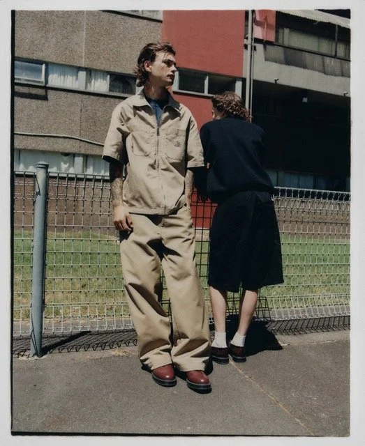 A young man with wavy hair and a young woman with curly hair standing by a fence outside a building. The young man is dressed in beige pants and a beige shirt, looking to the side. The young woman is leaning over the fence, with her back to the camer