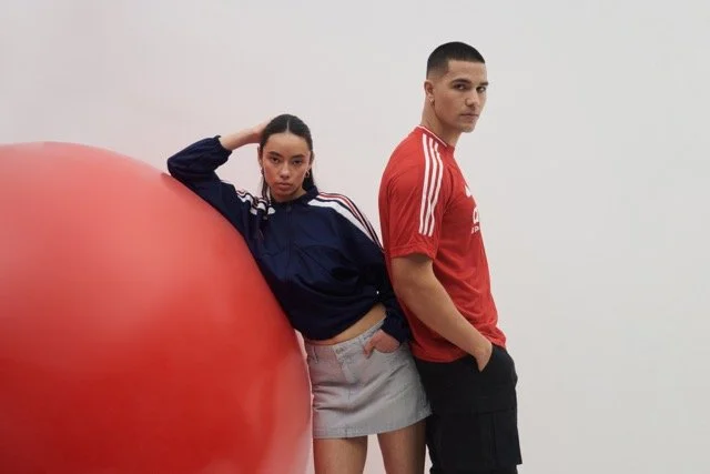 A young woman and a young man pose next to a large red exercise ball against a plain white wall. The woman is leaning against the ball with her arm on her head, wearing a navy blue jacket and a light gray skirt. The man stands beside her with his han