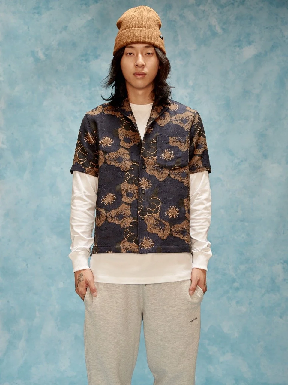 Young man with long hair wearing a tan beanie, floral patterned vest, white long-sleeve shirt underneath, and light gray sweatpants, standing against a blue painted backdrop.