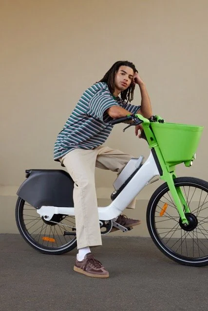 Young man sitting on a green electric bicycle, wearing a striped shirt and beige pants, with a neutral beige background.