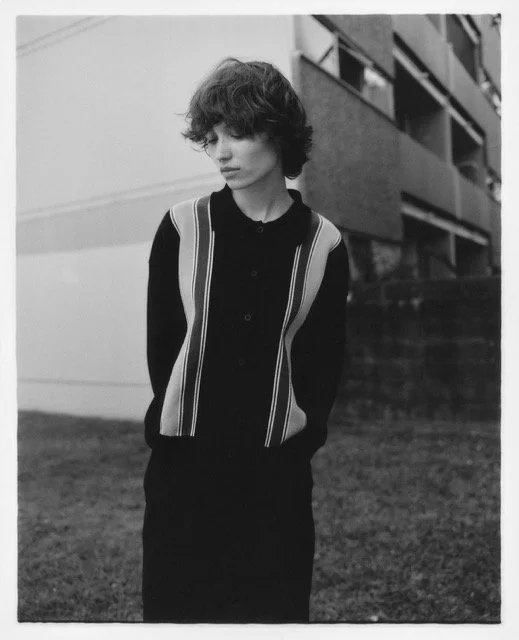 A young person with curly hair, wearing a black sweater with vertical stripes, standing outdoors in front of a modern building.