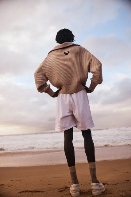 Person standing on a beach facing the ocean, wearing a tan sweater, pink shorts, and sneakers, with hands on hips and looking toward the sky.