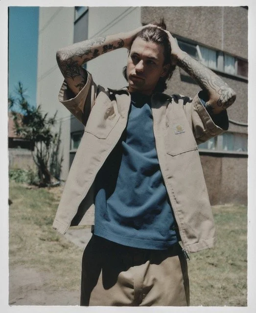 Young man standing outdoors in front of a building, adjusting his hair. He has tattoos on his arms and is wearing a beige jacket over a blue t-shirt.