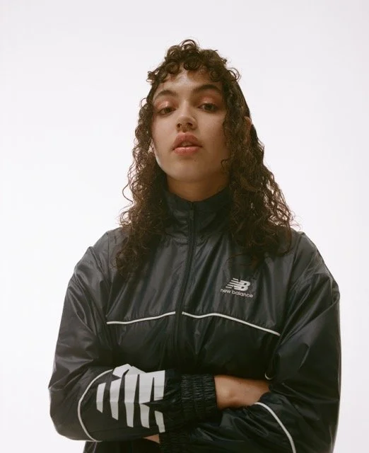 A young woman with curly hair wearing a black New Balance jacket with white accents, standing against a plain white background.