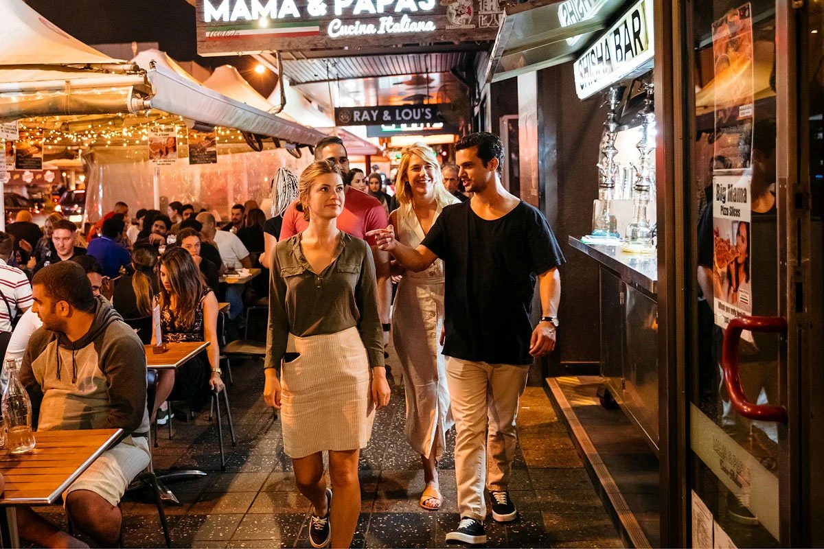 Group of young adults walking down a busy street with people dinning on the footpath during the evening.