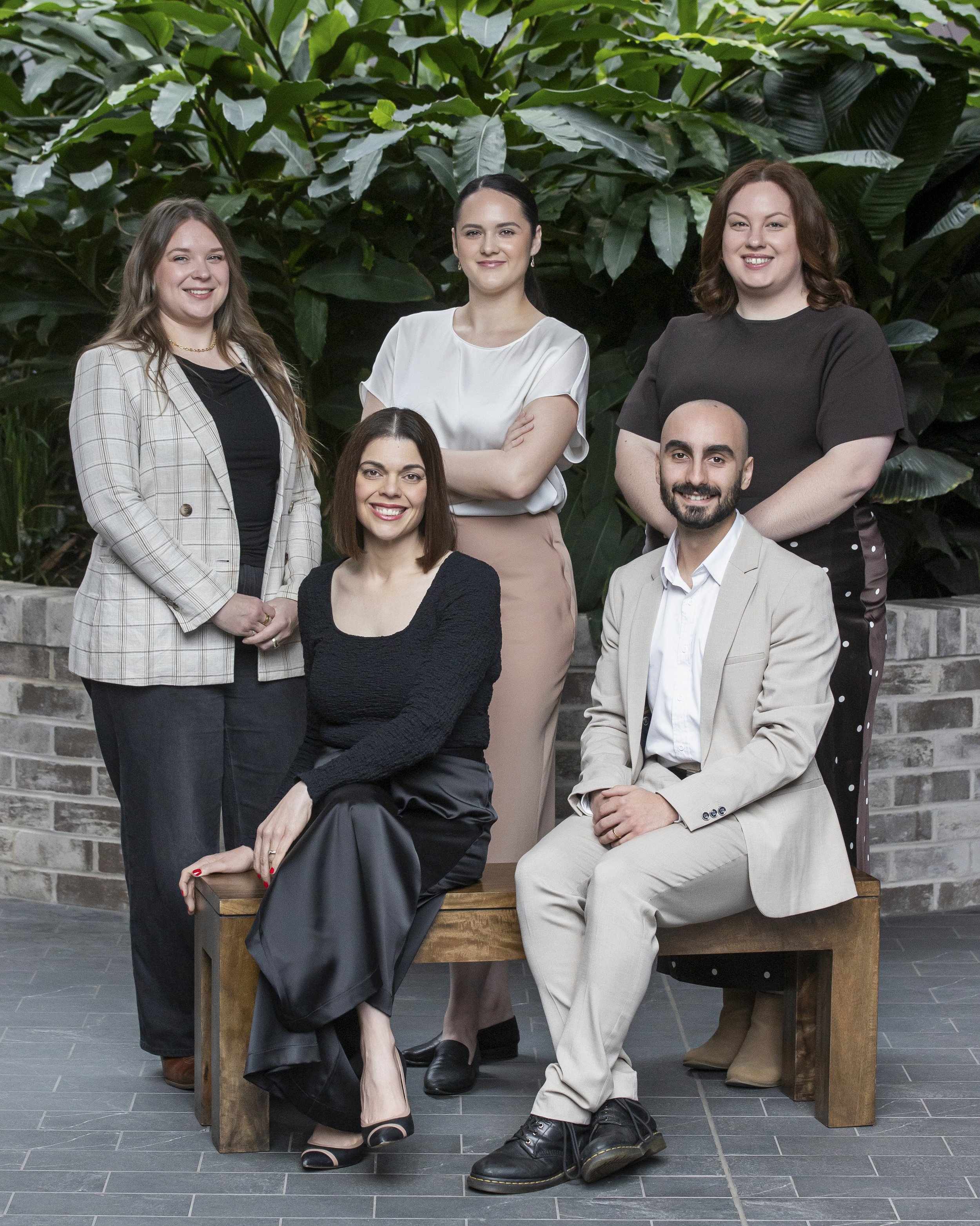 Group shot of Sparrowly Group team members with Beth, Morgan and Jackie standing in front of a brick wall and greenery,  and Giovanna and Federico sitting in front of them.