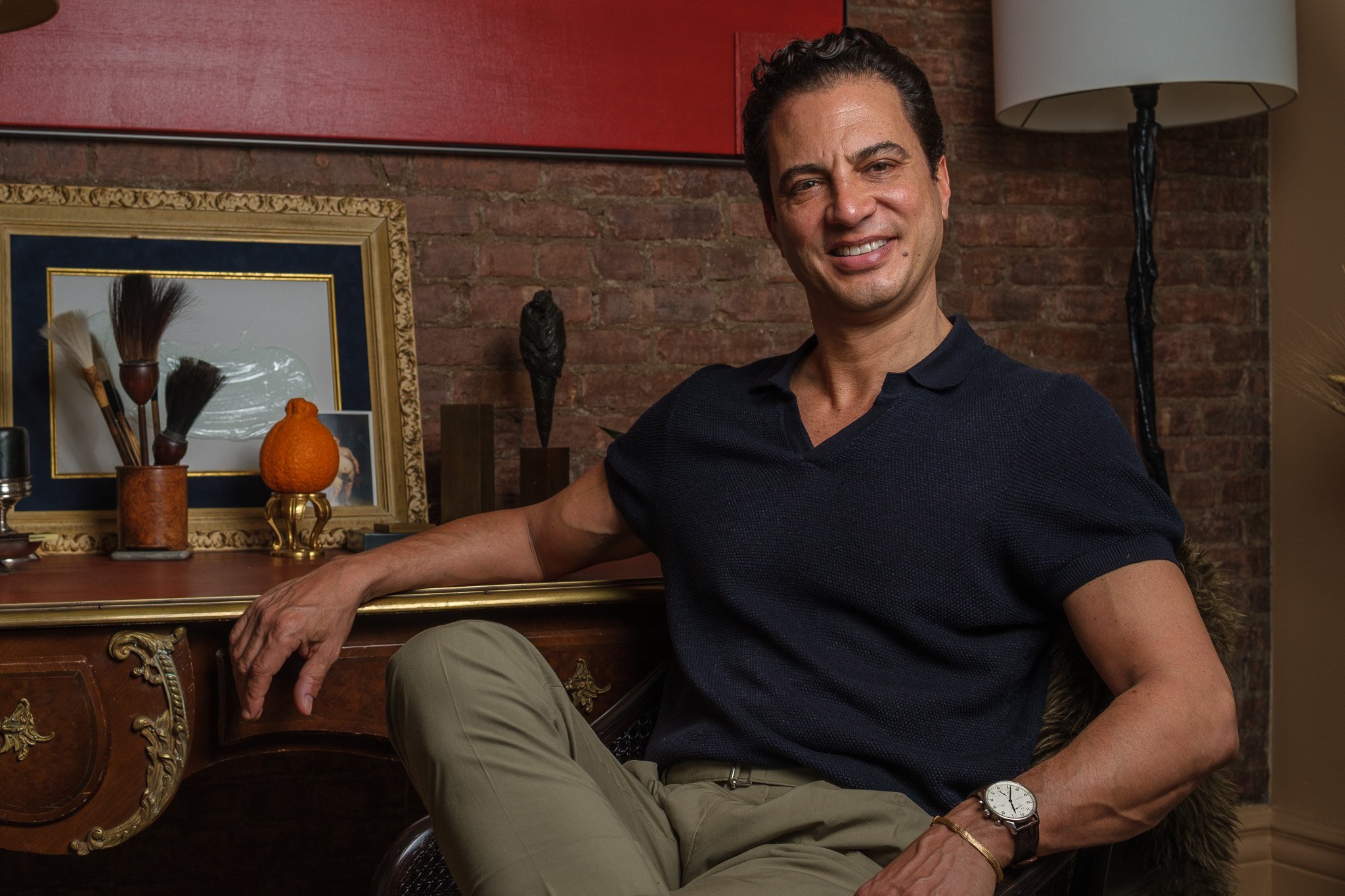 A man sitting in a room with a brick wall, leaning on an ornate wooden desk, smiling at the camera, wearing a dark short-sleeved shirt and beige pants, with a watch and bracelet on his left wrist.