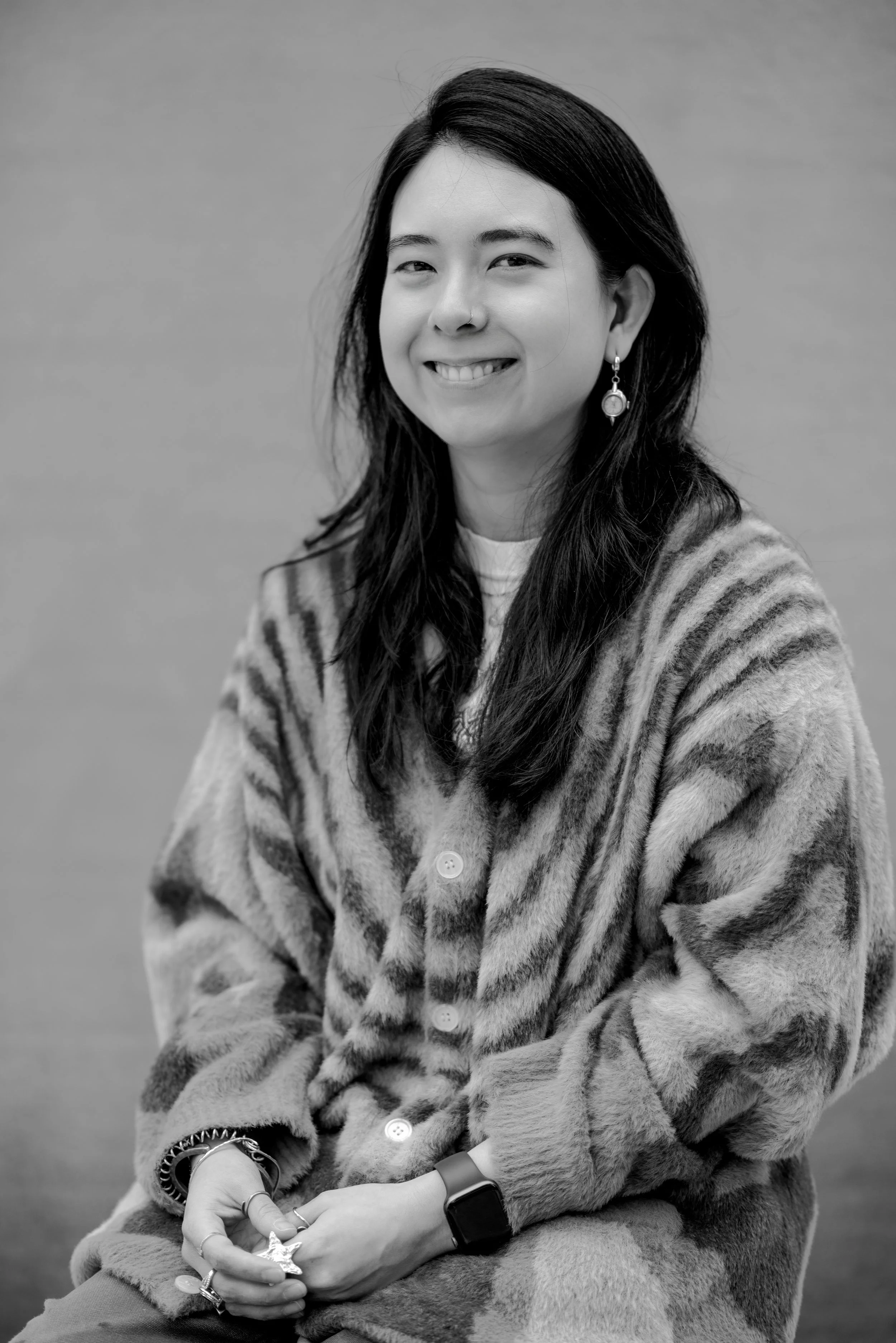 Portrait of a smiling woman with long dark hair, wearing earrings, a striped fuzzy button-up, and a smartwatch, sitting against a plain background.