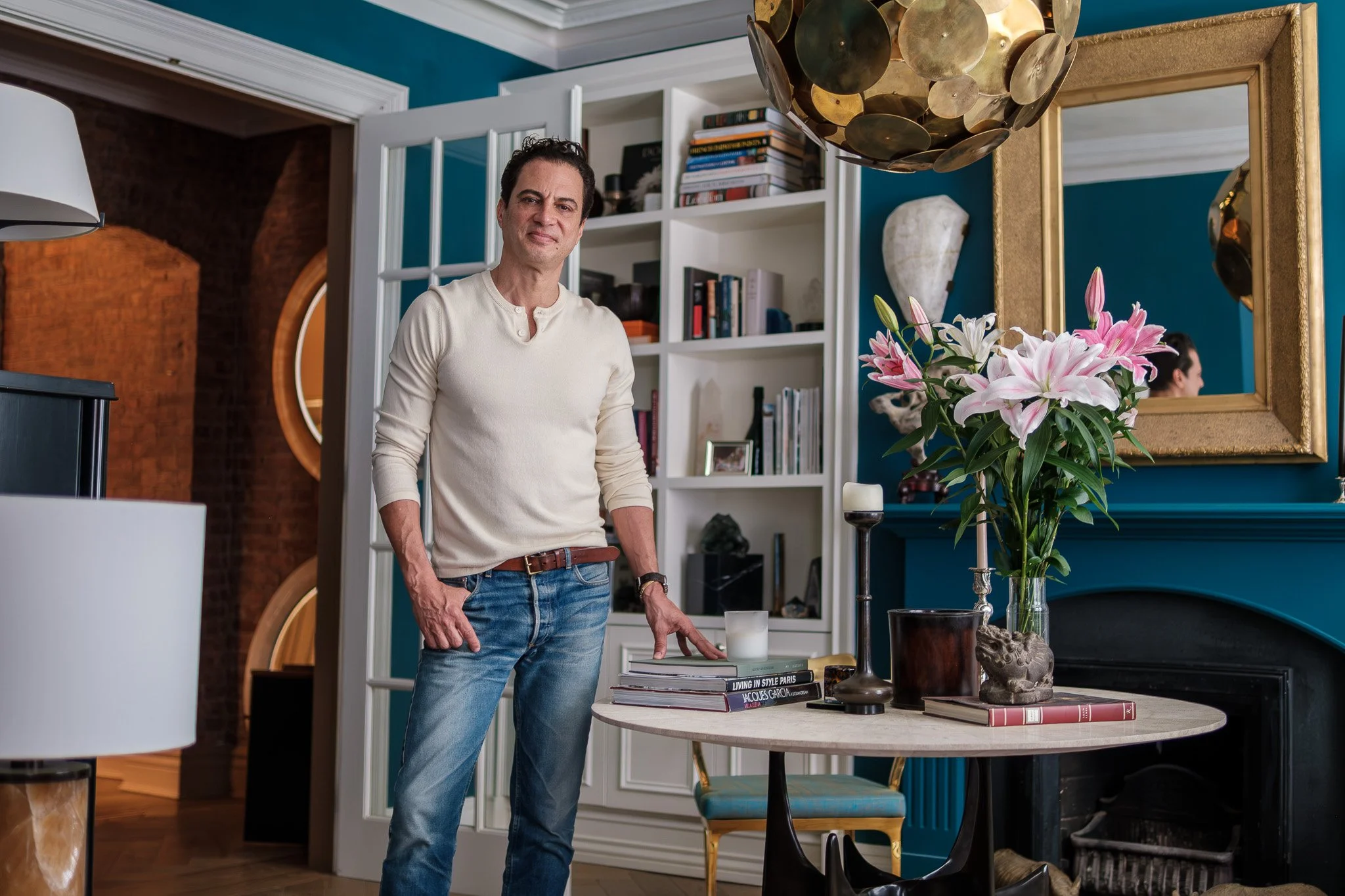 A man standing in a stylish living room with a blue wall, a white bookshelf, and a round table decorated with pink lilies, books, candles, and art pieces. He wears a white sweater and blue jeans.