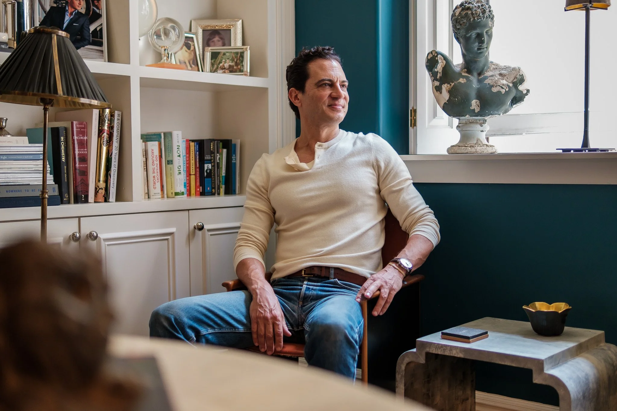 A man sitting in a room with bookshelves, a bust sculpture, and a window, looking to the side.