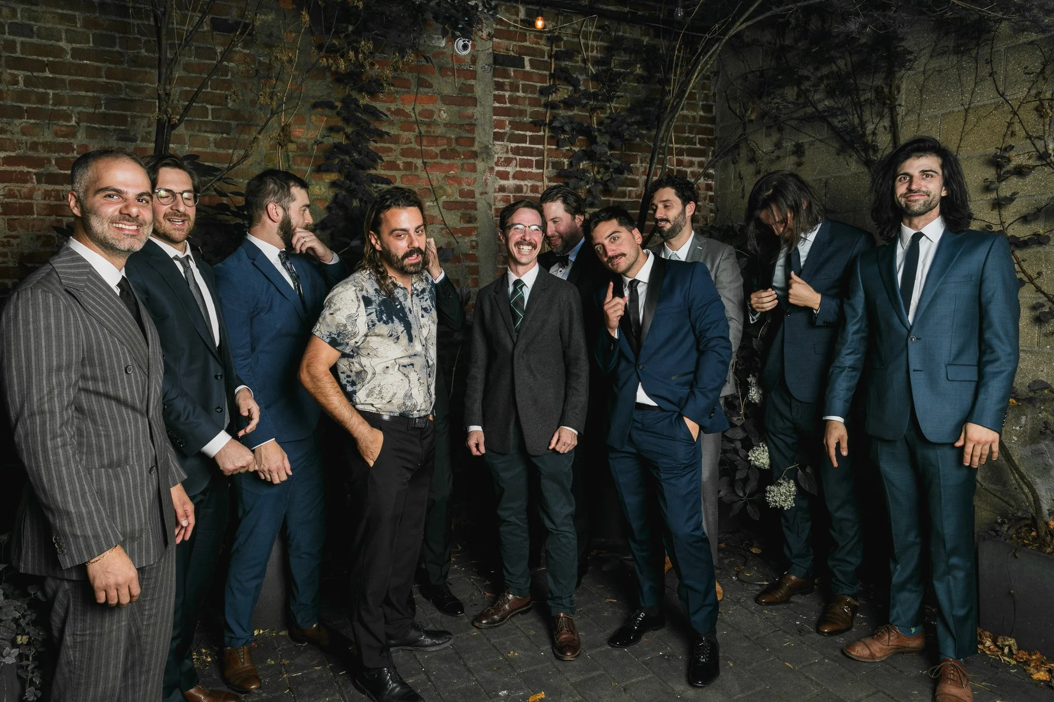 Group of eleven men dressed in formal and semi-formal attire standing against a brick wall adorned with plants, at night.