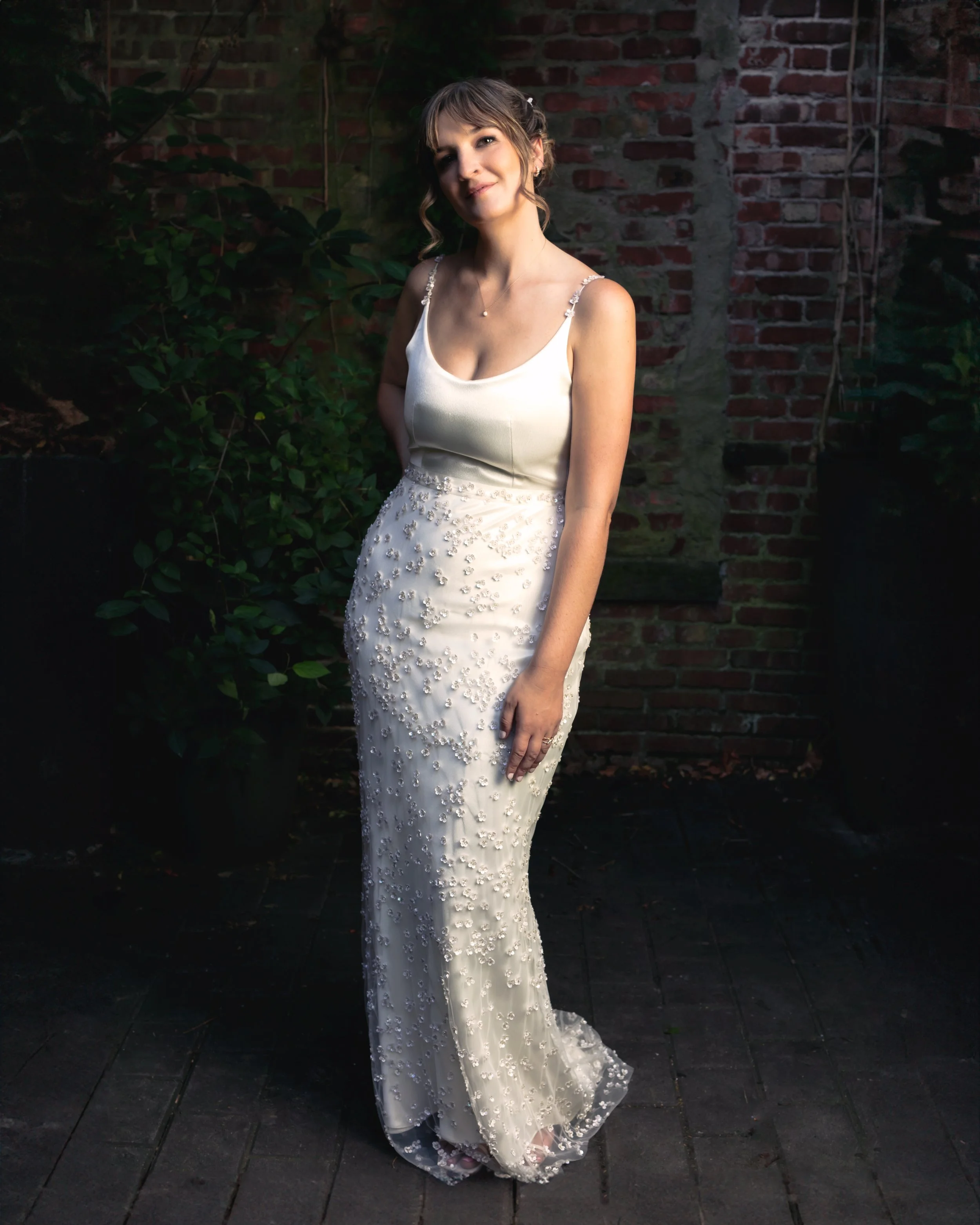 A woman in a cream wedding dress with beaded embellishments standing outdoors against a brick wall and greenery.