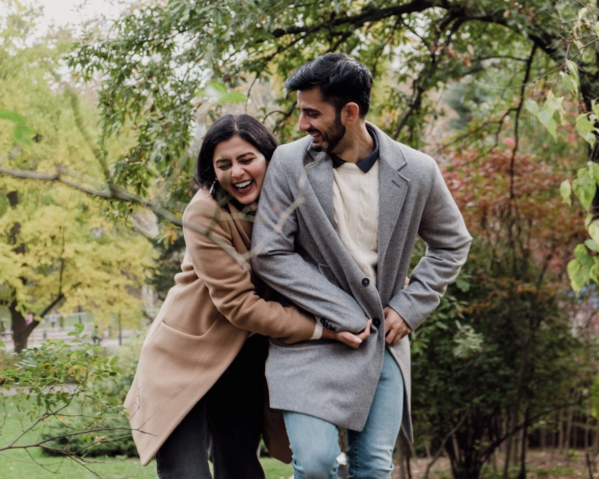 A happy couple laughing and playfully hugging outdoors in a park with autumn trees.