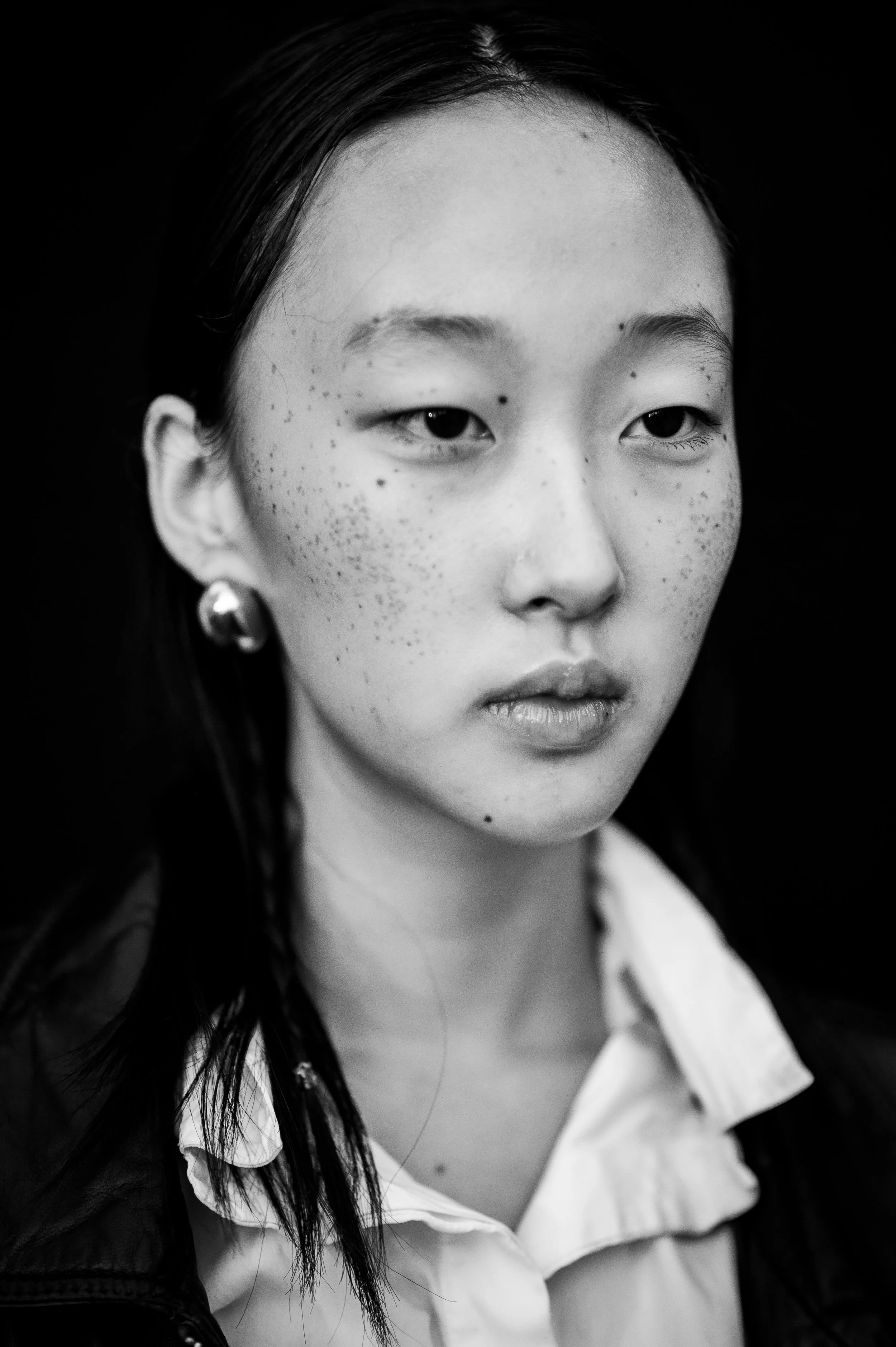 Close-up black and white portrait of a young woman with freckles, wearing earrings and a collared shirt, against a dark background.