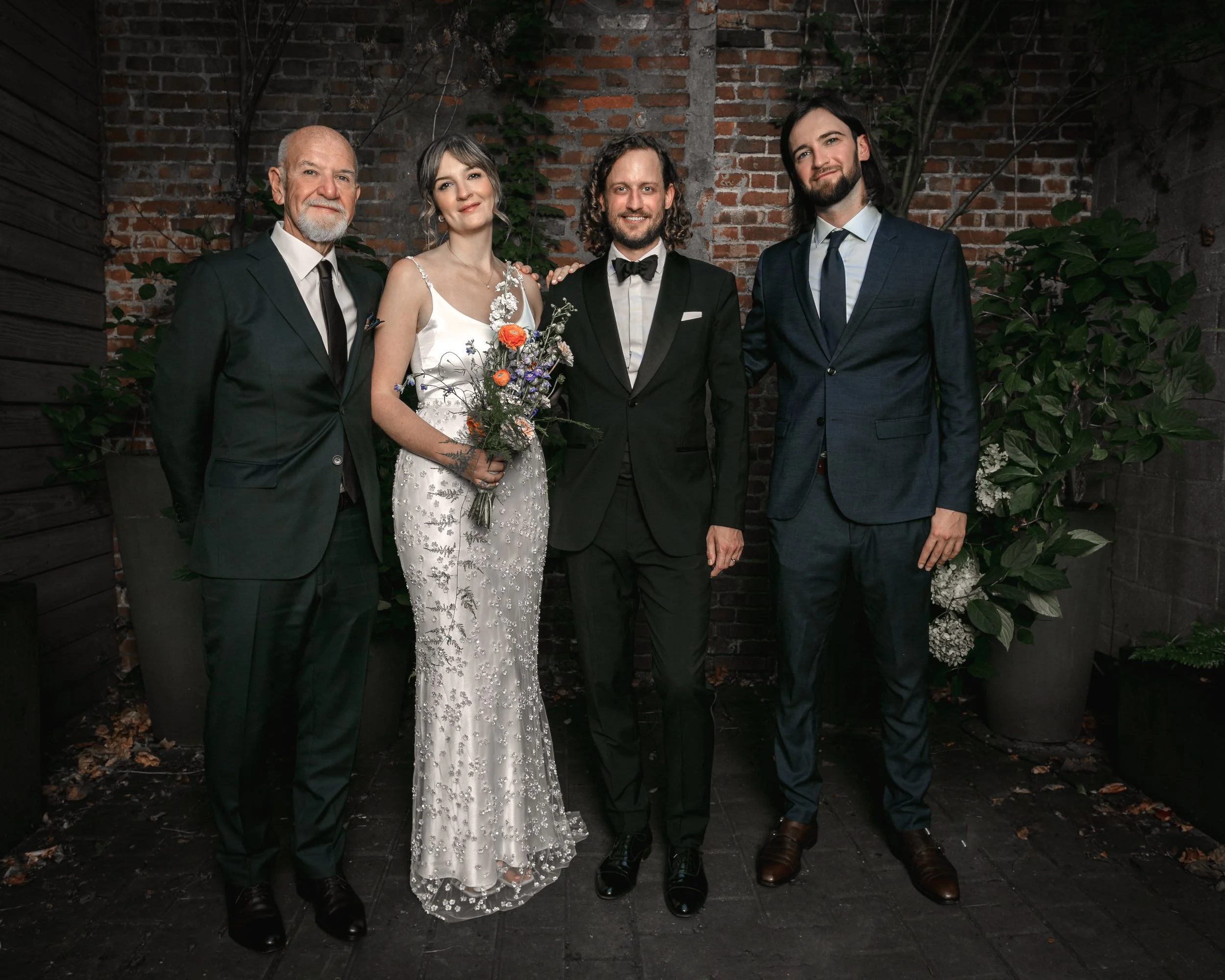 Group of five people in formal attire standing in front of a brick wall with greenery, posing for a photo at a celebration or event.