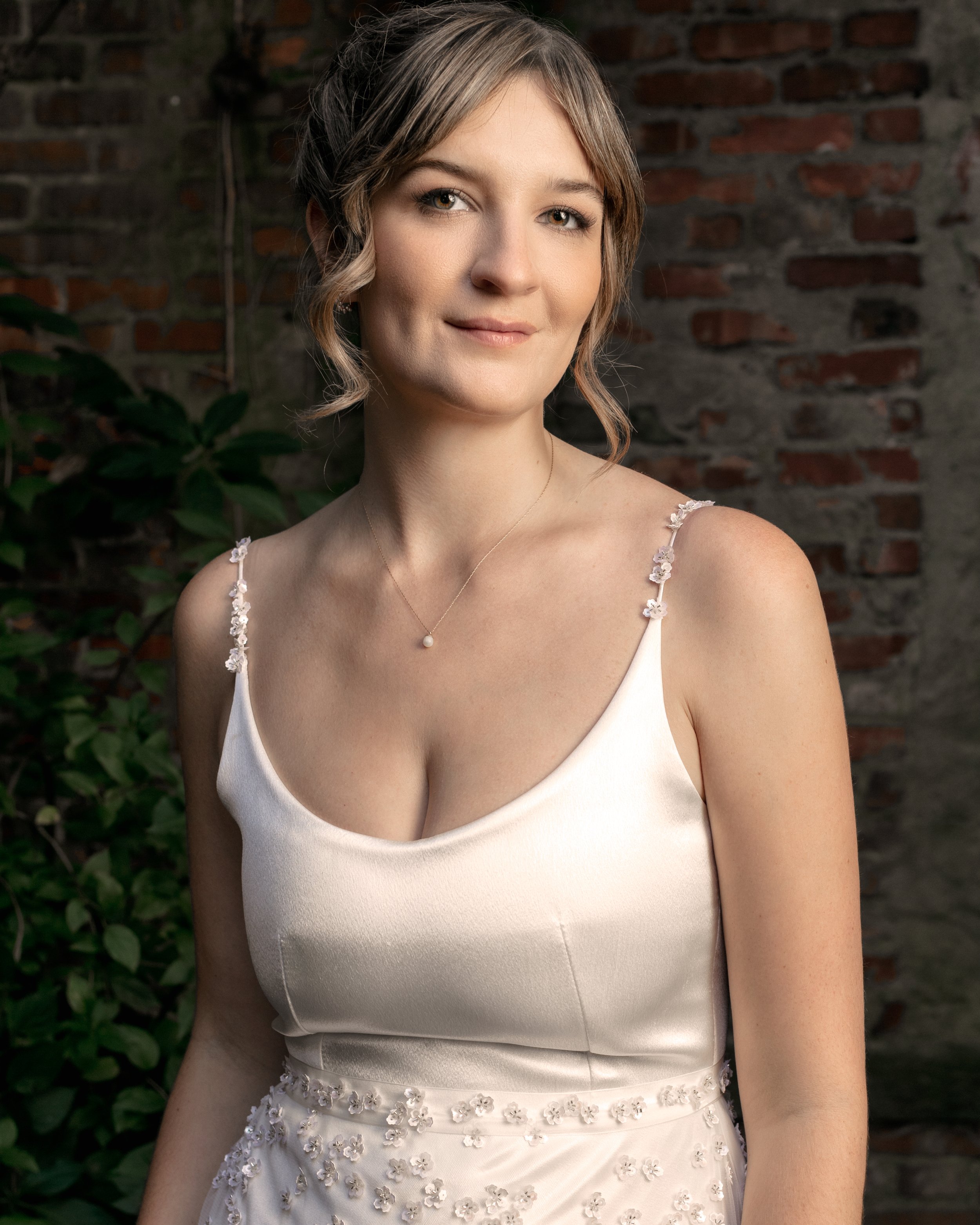 A young woman with shoulder-length light brown hair styled in loose waves and wearing a white dress with floral details on the straps and waist, standing in front of a brick wall with greenery to the side.