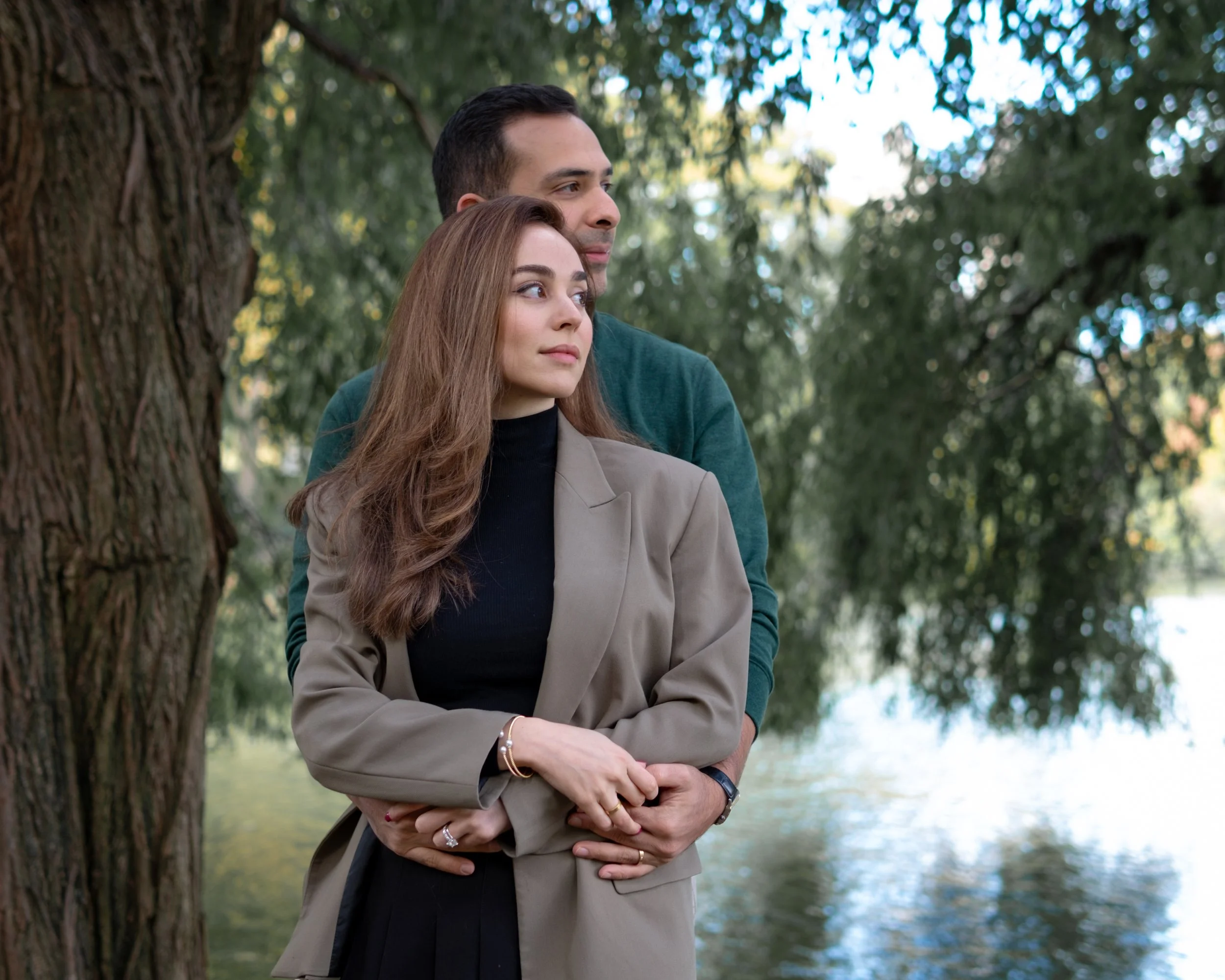 A couple hugging outdoors near a tree and a lake, with the woman in a beige coat and the man in a dark green sweater, both looking into the distance.