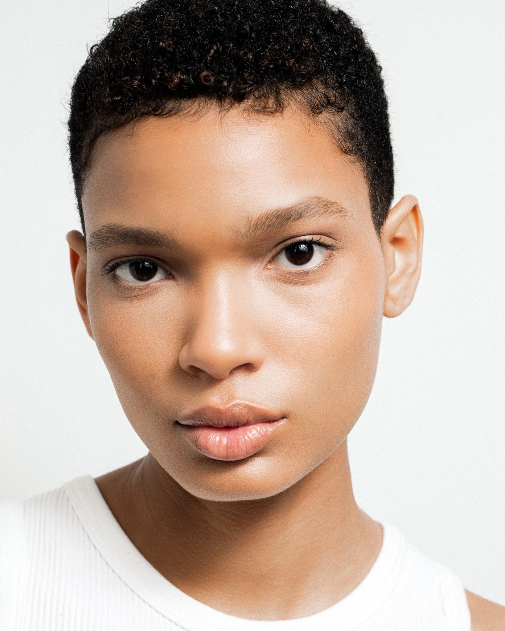 Close-up of a young woman with short curly hair and smooth skin, wearing a white sleeveless top, against a plain white background.